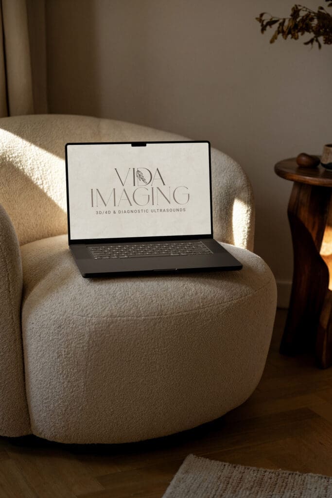 Vida Imaging Computer Mockup Laptop displaying the Vida Imaging logo on screen, styled on a cream accent chair in a warm, modern interior with natural light.