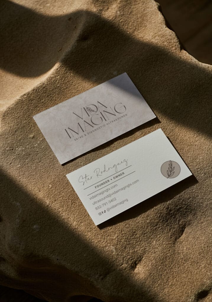 Vida Imaging Business Card Mockup Vida Imaging business cards featuring a minimalist beige logo and contact information displayed on textured stone in warm natural sunlight.